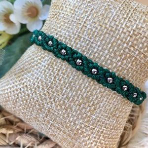 Op de afbeelding: Een groene gevlochten armband met zilveren kralen. De armband ligt op een jute achtergrond.