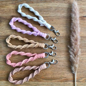 Op de afbeelding: Vijf macramé pols keychains in verschillende kleuren: gebroken wit, lavendel, beige, koraal en lichtbruin. Elke keychain heeft een gedraaid touwontwerp en een zilverkleurige sluiting. Ze liggen op een houten oppervlak.