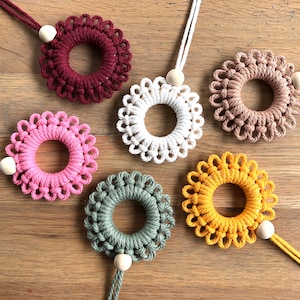 Op de afbeelding: Vijf macramé wandhangers in verschillende kleuren, elk met een houten kraal aan het einde van een touwtje. De kleuren zijn bordeauxrood, wit, beige, roze en geel.