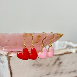 May include: A pair of gold heart-shaped earrings with red and pink enamel. The earrings are hanging from a pink and gold dish.
