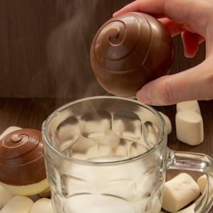 May include: A hand holding a chocolate sphere over a glass mug filled with milk and marshmallows. The chocolate sphere has a swirl design and is ready to be dropped into the mug to melt and create a hot chocolate drink.