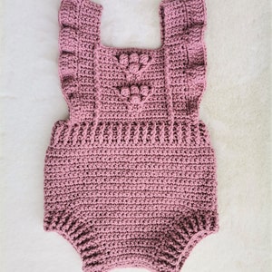 May include: A crocheted baby romper in a dusty rose color with a heart-shaped design and ruffled straps.