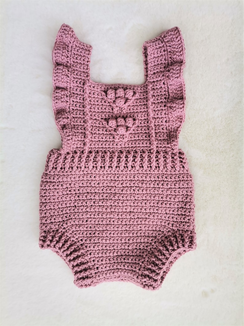May include: A crocheted baby romper in a dusty rose color with a heart-shaped design and ruffled straps.