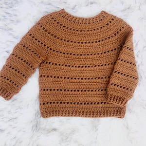 Puede incluir: Un suéter de crochet marrón con cuello redondo y mangas largas. El suéter presenta un diseño texturizado con filas horizontales de pequeños agujeros espaciados uniformemente. Los puños y el dobladillo son acanalados. El suéter se muestra sobre una superficie blanca y esponjosa.
