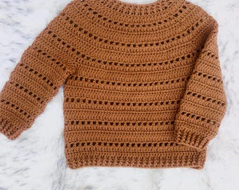 Può includere: Un maglione all'uncinetto marrone con scollo tondo e maniche lunghe. Il maglione presenta un design strutturato con file orizzontali di piccoli fori distanziati uniformemente. I polsini e l'orlo sono a coste. Il maglione è esposto su una superficie bianca e soffice.