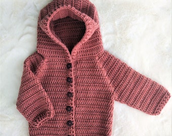 Pode incluir: Um casaco de crochê com capuz em cor rosa poeirento com botões marrons. O casaco tem um design simples e é feito com um fio macio.