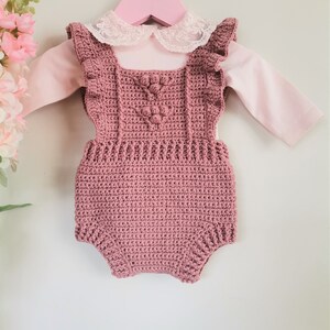 May include: A dusty rose crocheted baby romper with a white lace collar and ruffled straps. The romper is hanging on a pink hanger.