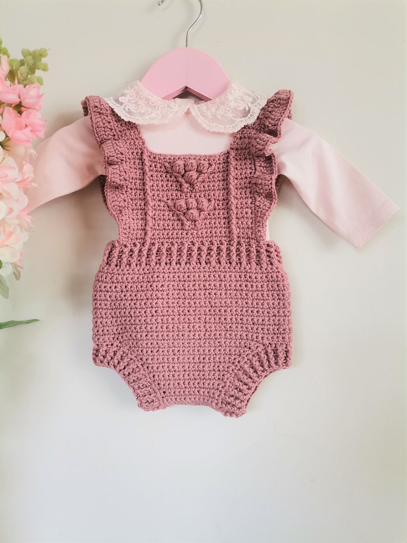 May include: A dusty rose crocheted baby romper with a white lace collar and ruffled straps. The romper is hanging on a pink hanger.