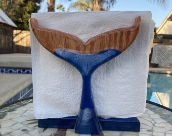May include: A wooden whale tail napkin holder. The top portion is natural wood, transitioning to a blue resin base. The holder is designed to hold a stack of white paper napkins, perfect for a nautical or coastal theme.
