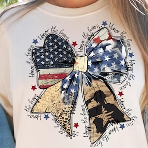 May include: Cream-colored t-shirt featuring a patriotic bow design. The bow incorporates the American flag, leopard print, and a silhouette of a soldier saluting. The words "Honor the brave" are written around the bow.