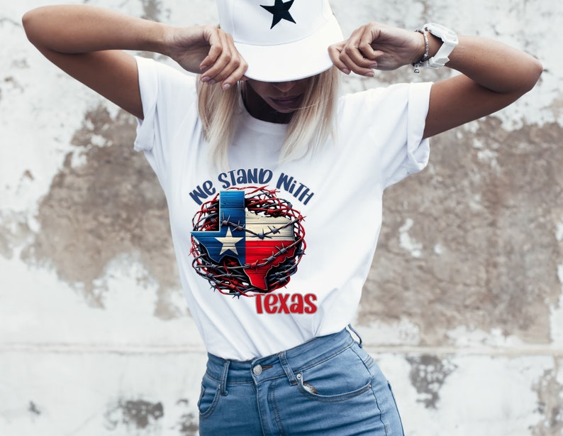 K&ouml;nnte beinhalten: Wei&szlig;es T-Shirt mit einer Grafik des Umrisses des Bundesstaates Texas in den Farben der amerikanischen Flagge, umgeben von Stacheldraht. Der Text "We Stand With Texas" ist auf das Shirt gedruckt.