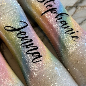May include: Three clear, iridescent glitter bottles with rainbow designs and personalized names. The names are "Stephanie", "Jenna", and "Jenna".
