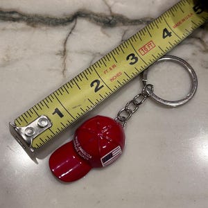 May include: A red baseball cap keychain with the text "MAKE AMERICA GREAT AGAIN" and an American flag design. The keychain is attached to a silver chain and ring. A yellow tape measure is in the background.