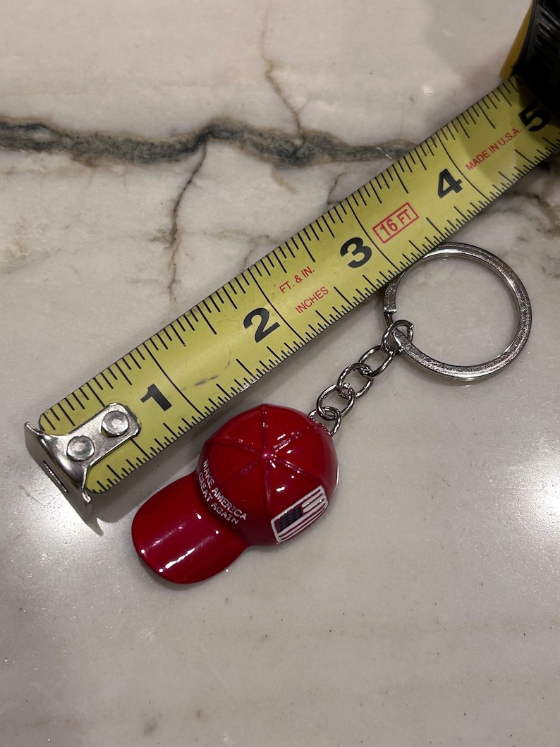 May include: A red baseball cap keychain with the text "MAKE AMERICA GREAT AGAIN" and an American flag design. The keychain is attached to a silver chain and ring. A yellow tape measure is in the background.