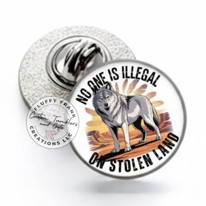 May include: A round pin with a wolf illustration and the text "NO ONE IS ILLEGAL ON STOLEN LAND." The pin has a silver backing and a white face with a gray wolf design. The pin is a custom creation.