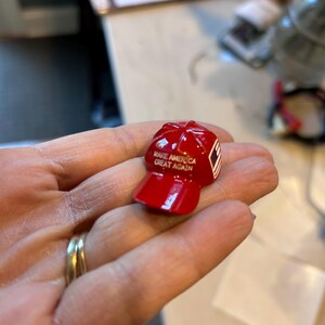May include: A small, glossy red baseball cap figurine. The cap has the text "MAKE AMERICA GREAT AGAIN" printed on the front. A small white and red design is on the side of the cap.
