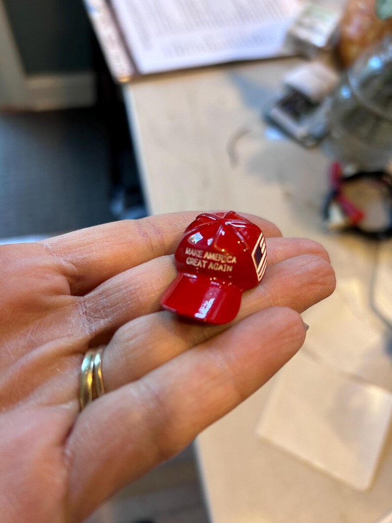 May include: A small, glossy red baseball cap figurine. The cap has the text "MAKE AMERICA GREAT AGAIN" printed on the front. A small white and red design is on the side of the cap.