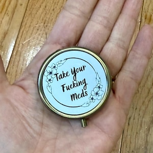 May include: A round, gold-toned compact mirror with the text "Take Your Fucking Meds" in black script. The text is surrounded by a floral wreath design. The mirror is held in a hand.