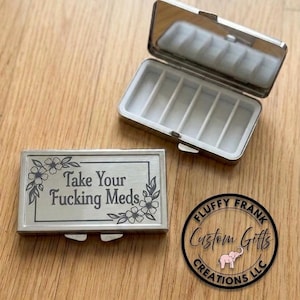 May include: A silver pill box with a hinged lid, open to reveal compartments. The box is engraved with the phrase "Take Your Fucking Meds" surrounded by floral designs. A circular logo is also visible.