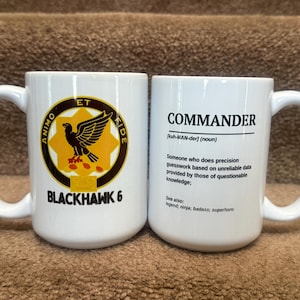 May include: Two white ceramic coffee mugs. One mug features a circular emblem with a black bird and the text "ANIMO ET RIDE BLACKHAWK 6". The other mug has the word "COMMANDER" and a definition printed on it.
