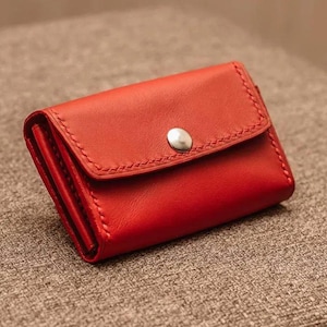 Peut inclure: Un petit portefeuille rectangulaire en cuir rouge doté d'un rabat repliable fixé par un bouton-pression en métal argenté. Le portefeuille présente des coutures visibles le long des bords du rabat et sur le corps principal de l'objet.