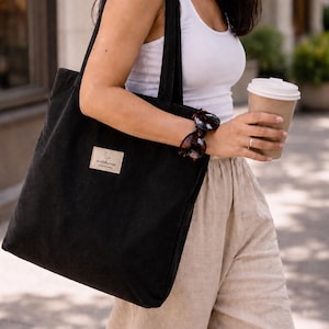 Puede incluir: Un bolso tote de pana negra con un parche rectangular de tela beige en la parte delantera. El parche muestra un pequeño icono de media luna sobre el texto "corduroyage" y las palabras "MADE IN INDIA" impresas en una fuente negra simple.