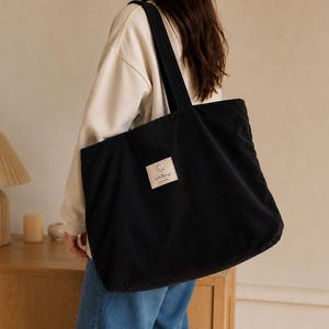 May include: A black corduroy tote bag with two shoulder straps and a rectangular beige fabric patch on the front. The patch features a small crescent moon illustration above the text "moonlight magic" and "made with love".