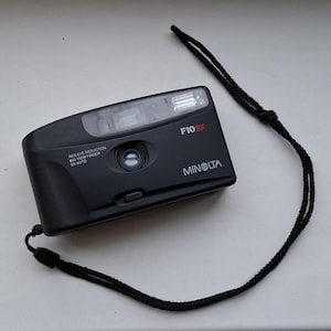 Könnte beinhalten: Eine schwarze Minolta F10BF Kompaktkamera mit roter Augenreduktion, großem Sucher und DX-Automatikfunktionen. Die Kamera hat eine schwarze Schlaufe.