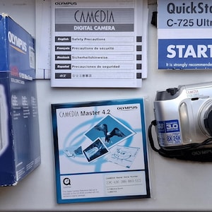 May include: An Olympus Camedia C-725 Ultra Zoom digital camera set including a blue retail box, a QuickStart Guide, a Camedia Master 4.2 software disc, a silver camera with a black lens cap and strap, and a green padded Olympus carrying case.