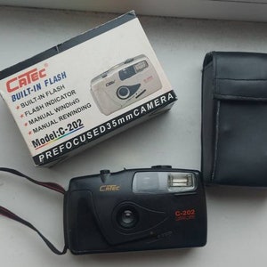 May include: A black 35mm film camera with a built-in flash. The camera has a black leather case and a strap. The box for the camera is white with black text that reads "Catec Built-in Flash Model: C-202 Prefocused 35mm Camera".