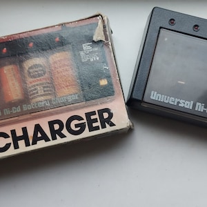 Puede incluir: Un cargador de batería universal Ni-Cd negro con una tapa de plástico transparente y cuatro luces indicadoras rojas. El cargador está sobre una superficie blanca junto a su embalaje original, que es marrón con texto blanco que dice "Cargador de batería universal de níquel-cadmio".