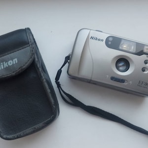 May include: A silver Nikon EF300 point and shoot camera with a black carrying case. The camera has a built-in flash and a lens cover.