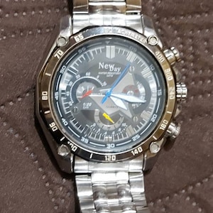 Op de afbeelding: Een zilverkleurig polshorloge met een zwarte wijzerplaat en een zilveren metalen band. Het horloge heeft een chronograaffunctie en een tachymeterschaal. Op de wijzerplaat van het horloge staan de woorden "NeWay" en "WATER RESISTANT 5ATM" gedrukt.