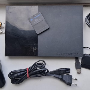 May include: A black Sony PlayStation 2 game console with a memory card, a black Sony DualShock controller, a black Trust Predator controller, a power cable, and an AV cable. The console features a rectangular design with a glossy and matte finish.