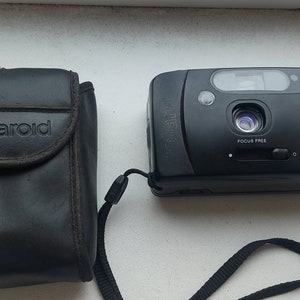 Op de afbeelding: Een zwarte Polaroid 2000FF camera met een zwarte leren tas en een band. De camera heeft een ingebouwde flitser en een 35 mm lens.