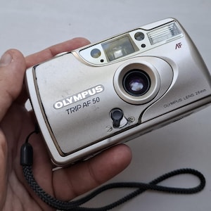 May include: A silver Olympus Trip AF 50 compact film camera featuring a 28mm lens, an autofocus system, and a built-in flash. The camera body includes a black wrist strap and clearly displays the brand name and model text on the front panel.