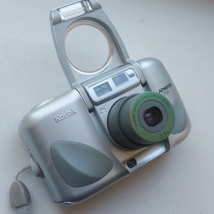 May include: Silver Kodak Advantix 750 camera with a green lens and a pop-up flash. The camera has a 2.5x zoom and a wrist strap. The Kodak logo is visible on the camera body.