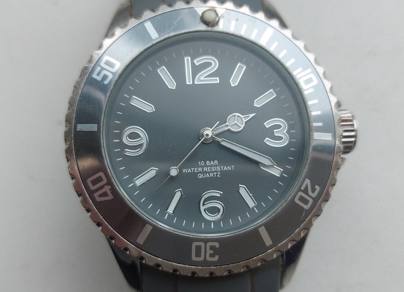 May include: A black and silver wristwatch with a black face and white numbers. The watch has a silver bezel with white markings and a black rubber band.