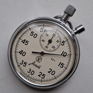 May include: A silver-toned mechanical stopwatch with a cream-colored face, black numerals, and two push buttons at the top. The dial features the text "Agat" in script, a small logo with "CCCP", "0,2 s", and "СДЕЛАНО В СССР" printed at the bottom.