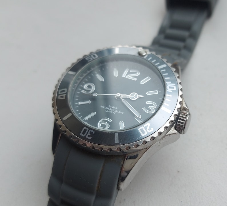 May include: A silver and black wristwatch with a blue face and white markings. The watch has a black rubber band and says "10 BAR WATER RESISTANT QUARTZ" on the face.