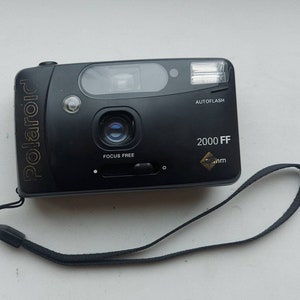 Op de afbeelding: Een zwarte Polaroid 2000 FF camera met een gouden logo en een zwarte band. De camera heeft een ingebouwde flitser en is gemarkeerd met "AUTOFLASH".