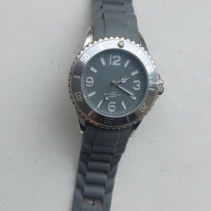 May include: A gray wristwatch with a silver face and black hands. The watch has a gray silicone band and a silver bezel with white markings. The watch face says "5 BAR WATER RESISTANT QUARTZ".