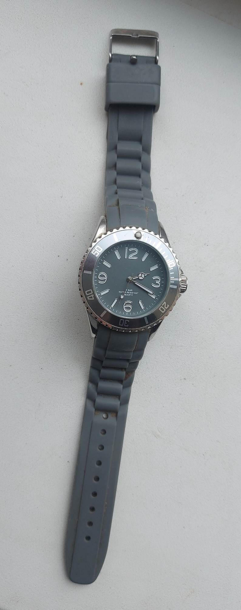 May include: A gray wristwatch with a silver face and black hands. The watch has a gray silicone band and a silver bezel with white markings. The watch face says "5 BAR WATER RESISTANT QUARTZ".