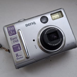 以下が含まれることがあります： 黒いレンズと内蔵フラッシュを備えた、シルバーのBenQ DC C30デジタルカメラ。3倍光学ズーム、3M解像度、8秒シャッターなどの機能があります。リストストラップと機能ラベルが含まれています。