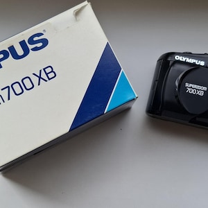 May include: An Olympus Superzoom 700 XB camera and its box. The camera is black with a lens cover and flash. The box is white with blue text and a blue and white angled design. The text on the box reads "Olympus Superzoom 700 XB Quartz Date."