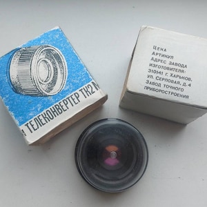 Puede incluir: Un objetivo de cámara vintage con cuerpo negro y elemento de vidrio transparente, acompañado de dos cajas de cartón. Una caja es azul y blanca con un diagrama, la otra es blanca con texto.