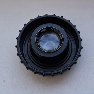 May include: A black lens with a silver ring and the text "AMAR PZO 58600 F:1.0-5cm" printed on it.