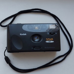 May include: A black Kodak Star AF point-and-shoot camera with a black strap. The camera has a built-in flash and red-eye reduction.