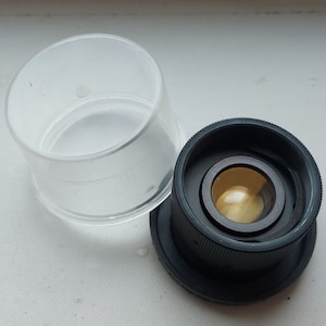 Puede incluir: Un adaptador de lente de cámara negro con un contenedor de almacenamiento de plástico transparente.