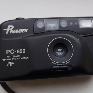 Puede incluir: Una cámara de película compacta negra Premier PC-850 con flash incorporado y enfoque automático. La cámara tiene una lente que dice "Lens Made in Japan".